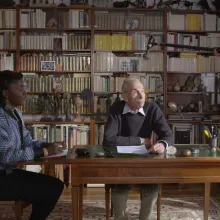 Alice Diop is a Senegalese-French woman wearing a multicolored jacket. She is sitting in a library, speaking to Pierre Bergounioux, an older white French male writer. They're both being filmed. From Alice Diop's 'Nous.' Photo by Sarah Blum. Courtesy of Mubi.