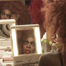 BeBe Zahara Benet is a Black drag queen preparing for a show in her dressing room in Minneapolis. She is wearing a very large brown wig and false eyelashes, while looking into a mirror. From Emily Branham’s ‘Being BeBe.’ Courtesy of the filmmaker.