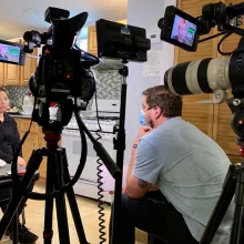 A light-skinned man wearing mask is sitting behind two large cinecameras on tripod, filming a Latino woman in the kitchen.