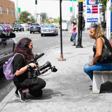 Margaret is a white woman wearing all black, filming Angela, a woman of color, wearing a black tank top and blue jeans. Behind-the-scenes shot from Margaret Byrne's 'Any Given Day.' Photo by Anjanli Pinto. 