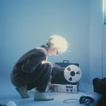 Maryanne Amacher is a white American composer and installation artist. She is wearing a gray jumpsuit and working in a room with musical equipment. From Lisa Rovner’s ‘Sisters with Transistors.’ Courtesy of Metrograph.