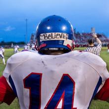 Back of a football player facing the field, his blue helmet has a horizontal tape with BELIEVE written with sharpie.
