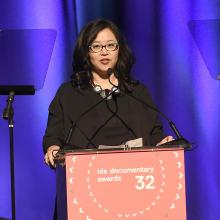 MacArthur Foundation's Kathy Im is an Asian American woman with shoulder-length black hair and glasses, wearing a black dress and standing at the podium at the IDA Documentary Awards.