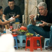 Anthony Bourdain, a white man with white hair, a black short and tattoos on his arms, sits on a sidewalk, eating a bowl of noodles. From  Morgan Neville's  'Roadrunner.' Courtesy of CNN / Focus Features