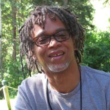 Lewis Erskine (1957-2021) is a middle-aged Black man with short dreads. He is wearing a grey t-shirt and black rimmed glasses. Image courtesy of Jean Tsien