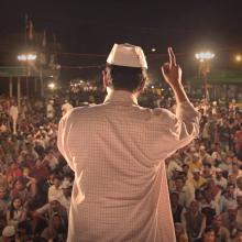 A man with brown skin in a white checkered shirt and white hat standing in front of a crowd. Still from 'An Insignificant Man' by Khushboo Ranka & Vinay Shukla. Courtesy of IFFLA. 