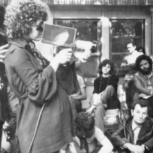 Black-and-white image of French feminist filmmaker Carole Roussoupolos, whose films are now streaming on “Another Screen.” In the photo, she is pregnant and is filming using a handheld camera, with people looking on. Courtesy of Another Screen; Centre Audiovisuel Simone de Beauvoir, Paris.