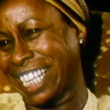 An image of Betty Carter, a Black female jazz singer. She is smiling, wearing a golden yellow blouse and a headband. From Michelle Parkerson’s “. . . But Then, She’s Betty Carter”. Courtesy of The Criterion Collection.