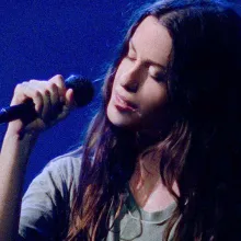 Singer Alanis Morissette, a white woman with long black hair, is performing on stage against a blue lit background; she is holding a microphone, and her eyes are closed, From Alison Kaynman’s ‘Jagged.’ Courtesy of TIFF.