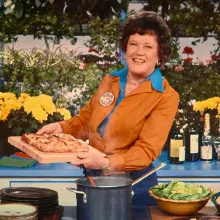 Julia Child is a white chef with short brown wavy hair. Here she is standing in her kitchen holding a tray of pastry. She is wearing a blue shirt underneath another brown one, and is smiling. Image from Julie Cohen and Betsy West’s ‘Julia.’ Courtesy of TIFF.