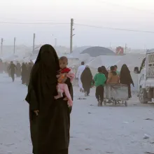  A woman in a burqa, her face covered, standing with a child on her lap. There are people walking behind her. Image from Hogir Hirori’s 'Sabaya.' Courtesy of Susan Norget Film Promotion.