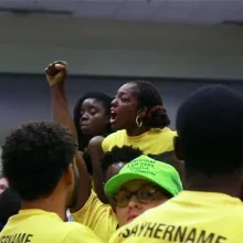 Caption: Janaé Bonsu is a young Black woman leading an activist gathering. She and her comrades are in yellow shirts that say “Say Her Name”. Image for Ashley O’Shay’s “Unapologetic”. Courtesy of David Magdael & Associates.