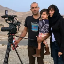Elizabeth Mirzaei and Gulistan Mirzaei standing in an Afghanistan desert with a camera, in 2019. Gulistan is carrying their daughter, Maryam. Elizabeth is wearing a headscarf, Gulistan is wearing a black t shirt and glasses, Maryam is wearing a striped shirt and floral pants. Image courtesy of the authors.