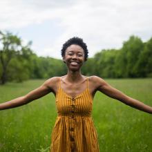 Elodie Edjang, a Black woman with short hair and a yellow dress stands in a grass field.