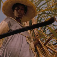 A Black woman farmworker stands against a sugarcane plant with a sickle in her hand in Haile Gerima’s ‘Sankofa’