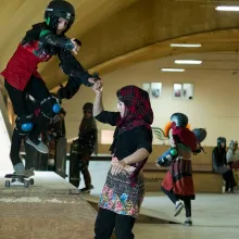 A young Afghani girl in a checkered headscarf helps her friend on a skateboarding ramp. Her friend is wearing skateboarding gear. They’re inside a training room with other young skateboarders. Image from Carol Dysinger’s ‘Learning to Skateboard in a Warzone (If You're a Girl).’ Courtesy of the filmmaking team.