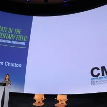 Caty Borum Chattoo, a white woman standing behind a podium, presenting the 2018 State of the Documentary Field Report. A presentation screen behind her. Courtesy of CMSI