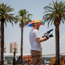 John Wilson is a white male filmmaker wearing a colorful sun umbrella around his head. He is holding a video camera with palm trees in the background. From ‘How To with John Wilson.’ Image courtesy of Joe Buglewicz/HBO.