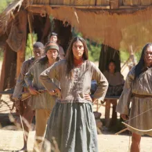 A group of 19th century Seminole tribespeople, wearing long-sleeved tunics and armed with bows & arrows, guard their dwelling.