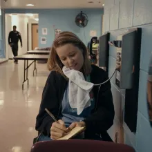 A white woman with shoulder-length red hair talks on a pay phone in a hallway of a correctional facility. From Jennifer Redfearn and Tim Metzger’s ‘Apart,’ which airs February 21 on ‘Independent Lens.’ Courtesy of the filmmakers.