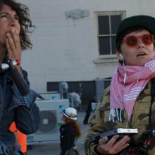 Cinematographer Jenni Morello, the author of this essay, is at left holding her camera in her right arm, her left hand is covering her mouth, and she's looking to her upper left. Director Ana Veselic is at right, holding a sound monitor in her right hand and looking and pointing to her upper left. 