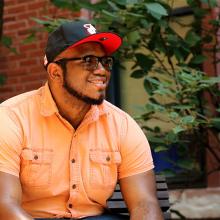   Headshot of a smiling Black adult man sitting on a bench in a black and red hat, glasses, and an orange button-up shirt.