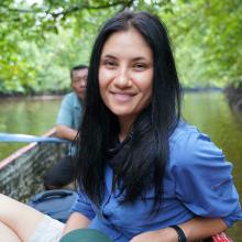 Allison Hanes, a woman with light skin and long black hair, sits in a small boat on a river. An older man with brown skin and dark hair sits behind her. Image courtesy of Hanes.
