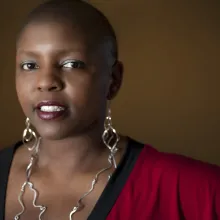 Yoruba Richen is an African American woman. She has a shaved head and is sporting large abstract dangled metal earrings that reaches her chest. She is wearing a red V-neck top with contrasting black neck band.