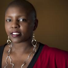 Yoruba Richen is an African American woman. She has a shaved head and is sporting large abstract dangled metal earrings that reaches her chest. She is wearing a red V-neck top with contrasting black neck band.