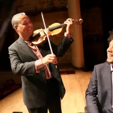 One Latino man, wearing a grey suit jacket, plays the violin, while the other, wearing a black suit jacket, watches and smiles.