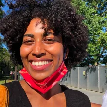 A Black woman with a huge smile and short curly hair is seen wearing a red mask on half of her face in a green and luscious park.