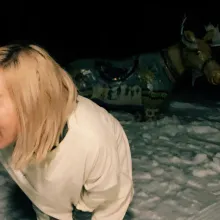 Woman of Korean-descent, she has a big smile and blonde hair to her shoulders. She is looking to her right, in the snow at night with a moose sculpture behind her.