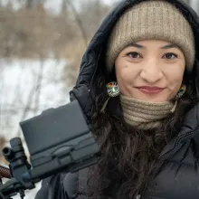 Tsanavi Spoonhunter is a Northern Paiute and Northern Arapaho woman with long, curly dark brown hair and eyes smiling into the camera.