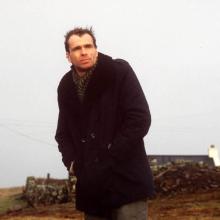 A man in his 30s, wearing a black overcoat, stands on a country road