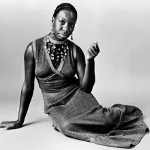 Nina Simone is sitting on the floor with her right arm propping her up. She is wearing a maxi knit dress, large decorative necklace.