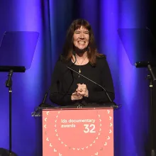 Dianne Weyermann speaking at a podium at the 2016 IDA Documentary Awards. She is wearing black and has her hair untied.