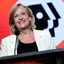 PBS President and CEO Paula Kerger at a podium, wearing a white blazer over a black blouse with the PBS logo behind her.