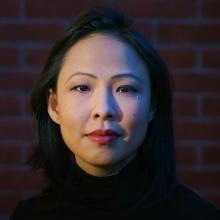 An Asian woman of Malaysian Chinese origin with shoulder-length black hair. She is wearing a black turtleneck & in front of a brick wall.