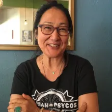 Rea Tajiri, standing in front of a mirror, wearing a black t-shirt, glasses, and a necklace with her hair tied up, smiling.