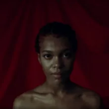 A Black woman with short curly hair stands in front of a red background, bare-shouldered and staring at the camera. From Juliana Kasumu’s Baby Bangz: Black Power in Hair.