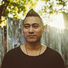 PJ Raval is an Asian man with a short mohawk and high fade, dressed in a round-neck black tee. He is in front of a fence next to a tree.