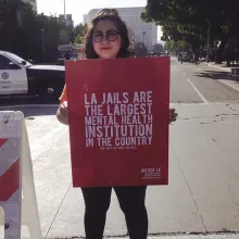 ​A woman wearing a hat and glasses, with shoulder-length hair, stands on a street and holds a sign that reads "LA Jails are the largest mental health institutions in the country." From Kenneth Paul Rosenberg’s 'Bedlam.' Courtesy of 'Independent Lens.'