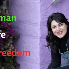 Headshot of a young white woman with long black hair and a black top in front of a purple wall. On the left side of the picture, we see Woman (in green), Life (in white), and Freedom (in red).