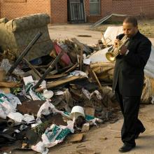 Film still from Spike Lee's 'When the Leeves Broke: A Requiem in Four Acts'., featuring a Black man in a suit playing the trumpet while walking next to a trash-filled sidewalk.  