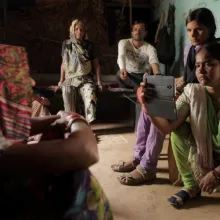 At right, A reporter from an India-based digital platform is kneeling while interviewing an Indian woman who is seated at left, in her living space with her family. The reporter is filming with a smartphone in her right hand , nd is wearing an emerald green sari