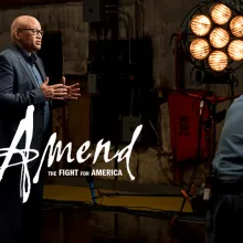 Larry Wilmore, an African American male film director, is wearing a black blazer over a blue shirt, talking to someone off screen on a film set. 