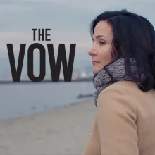 A light-skinned woman, wearing a grey scarf and cream coat, is looking out on a beach
