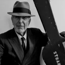 An image from Hallelujah: Leonard Cohen, A Journey, A Song, Leonard Cohen is wearing a fedora and holding a guitar in a black & white photo. 