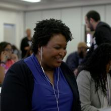 A Black woman in blue shirt and black cardigan, listening to her headphones sitting down