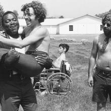 Teenagers with disability at summer camp. A Black man is holding a light-skinned man with curly hair.